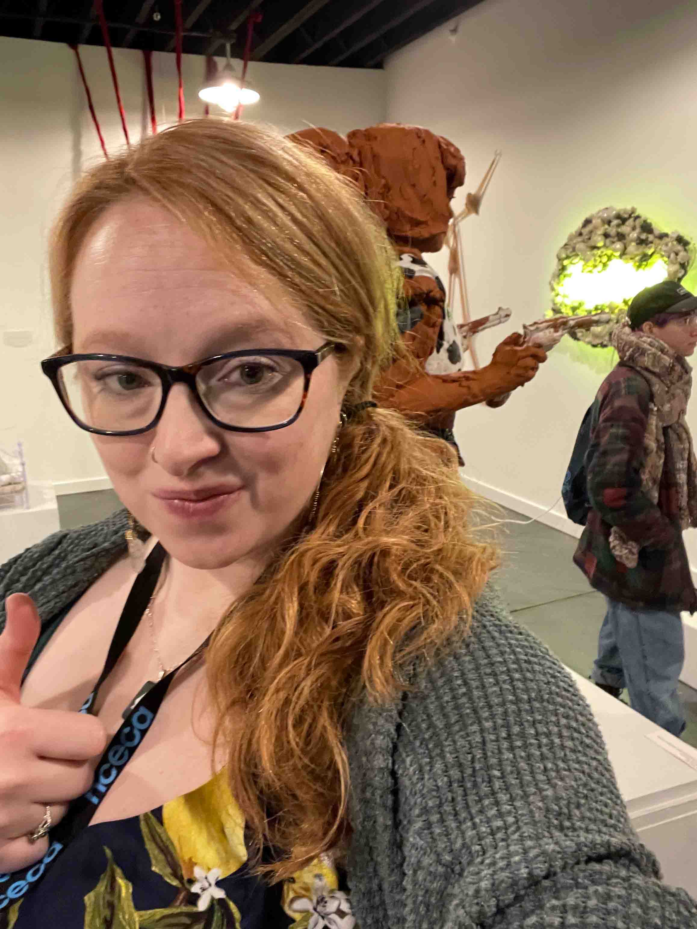 A woman with her thumbs up taking a picture smiling in an art gallery