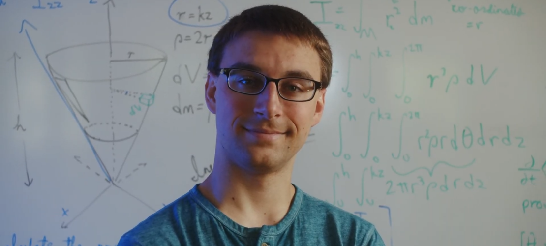 a student faces us as he stands in front of a whiteboard covered with equations