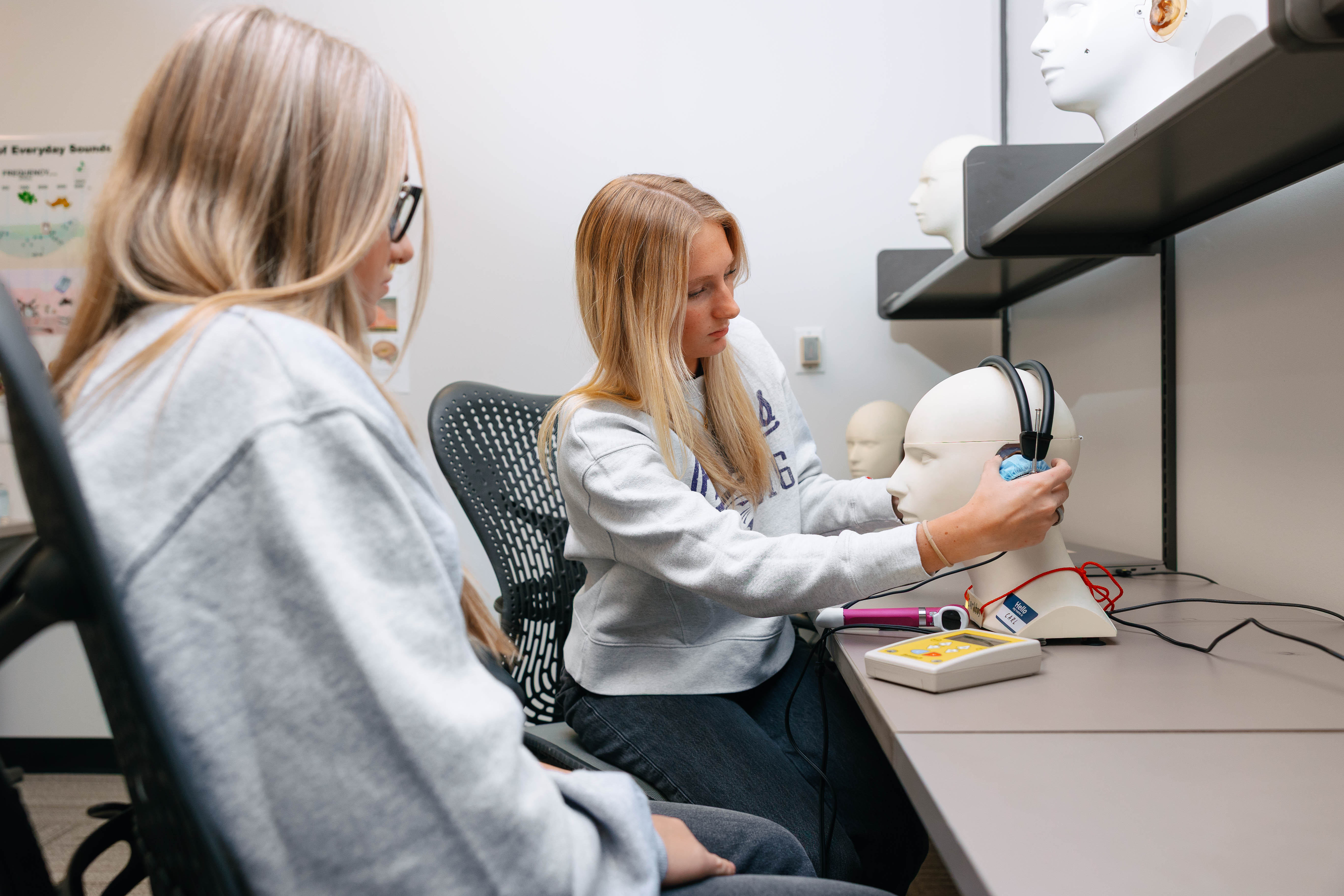 Two students sit at a desk in a lab setting as one adjusts headphones on a mannequin head connected to testing equipment, while the other observes.
