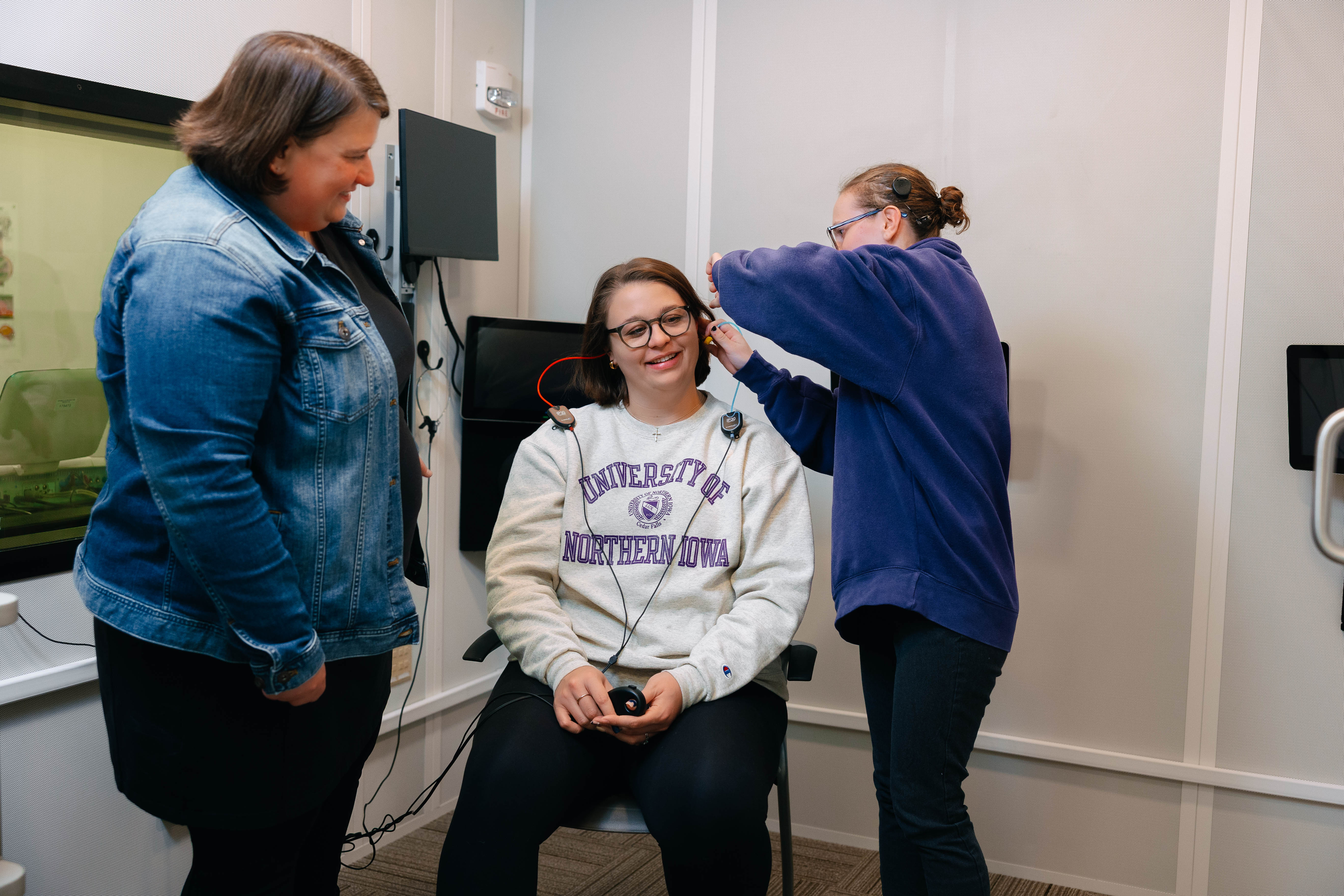 A woman sits in a chair wearing headphones connected to small monitoring devices while another woman adjusts the earpiece and a third woman stands nearby observing in a clinical testing room.