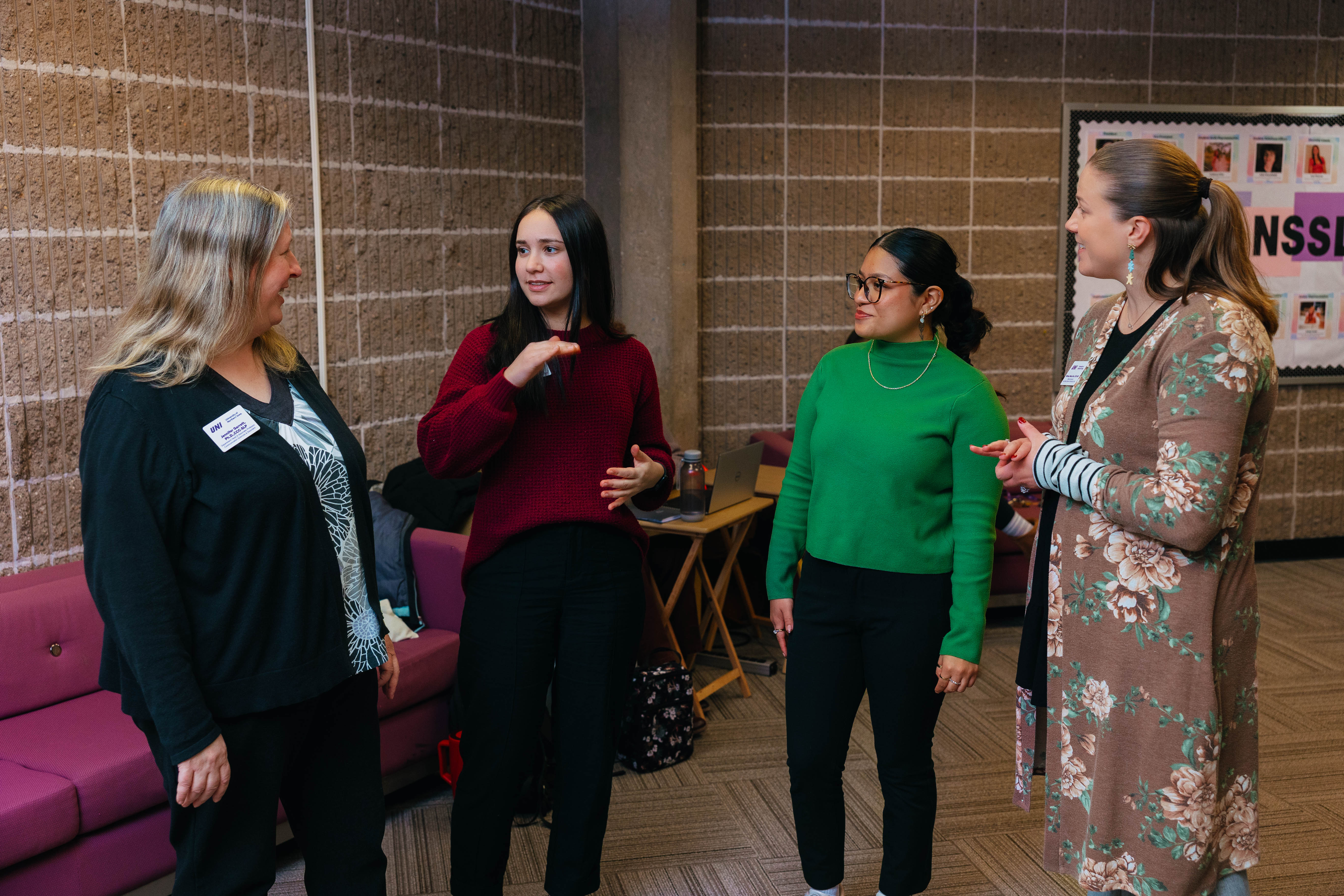 Four women stand in a lobby space talking together, smiling and gesturing during a conversation, with a brick wall and bulletin board in the background.