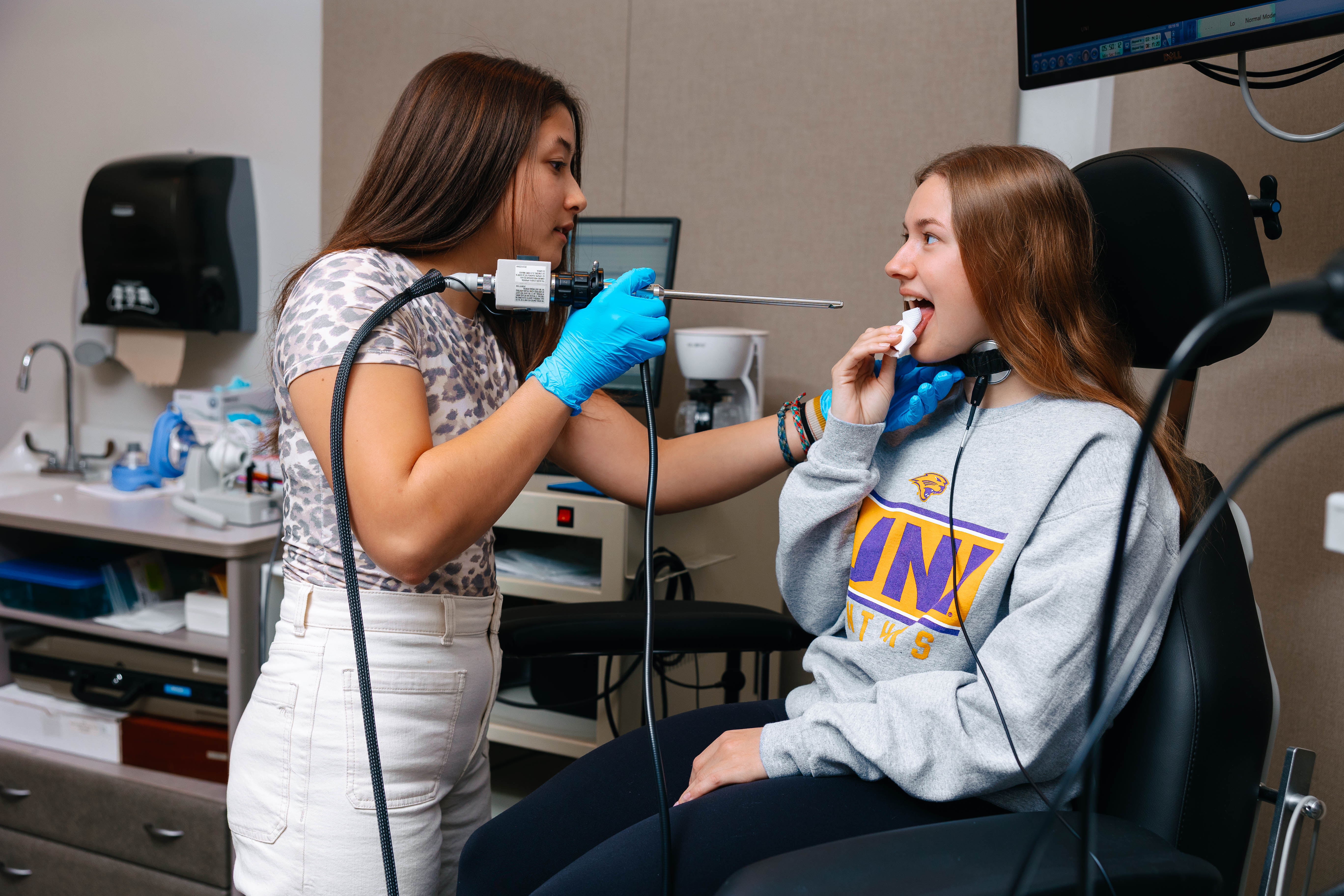 A clinician wearing gloves uses a small scope to examine a seated student’s throat in a clinical room, while a monitor displays a close-up image of the vocal cords during the procedure.