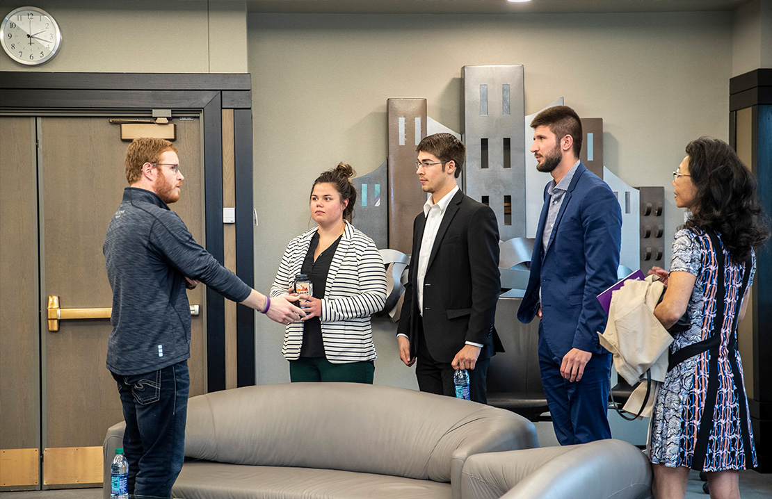 A small group of students and faculty stand in conversation inside a modern campus lounge, with several people dressed in business or business-casual attire, suggesting a professional or networking setting.