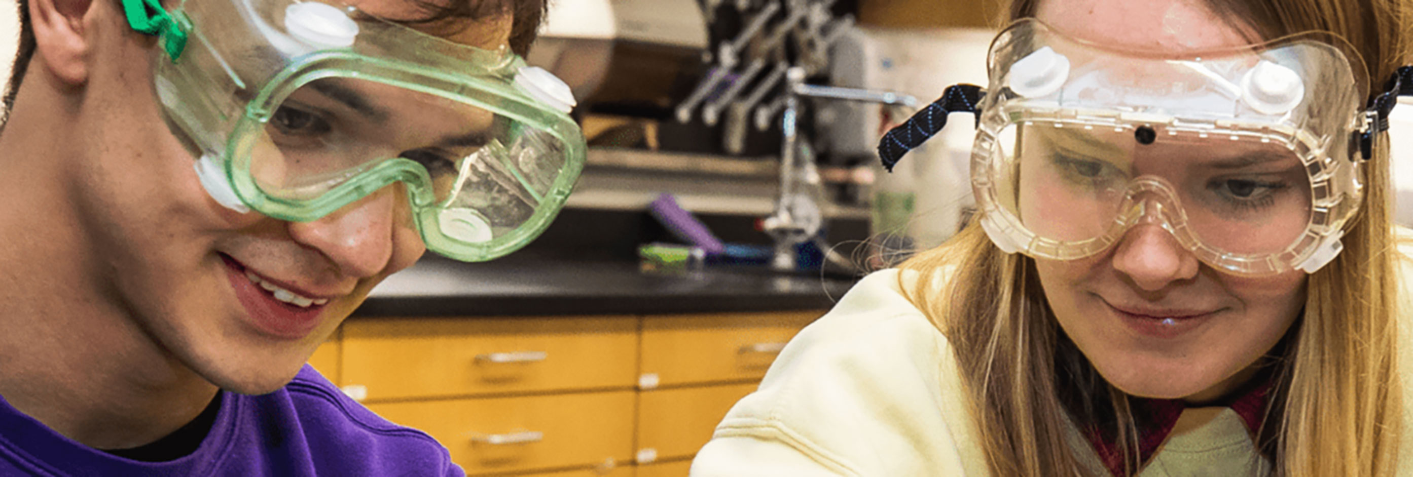 two students in safety goggles work with lab equipment