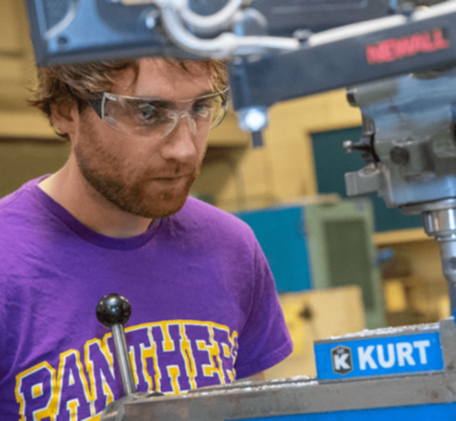 Student in a Panthers shirt and safety goggles inside a shop.