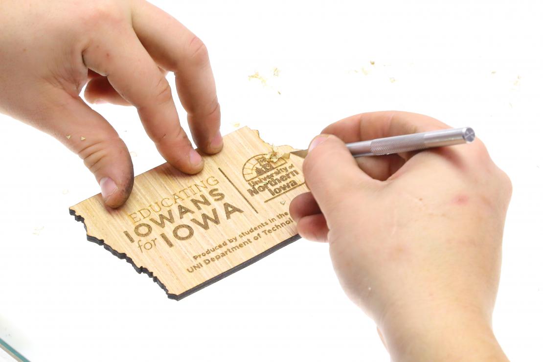 we see a students hands working with a scalpel to trim the excess of the edges of a small wooden cutout of Iowa