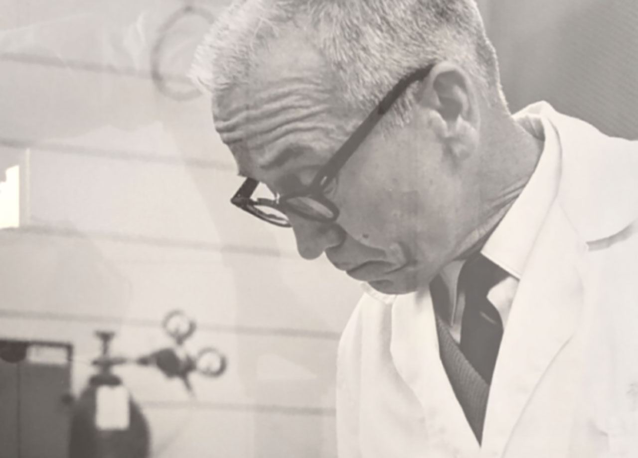 Close-up of Dr. Leland Wilson in a chemistry lab, wearing a white lab coat and black glasses, looking down at his work.